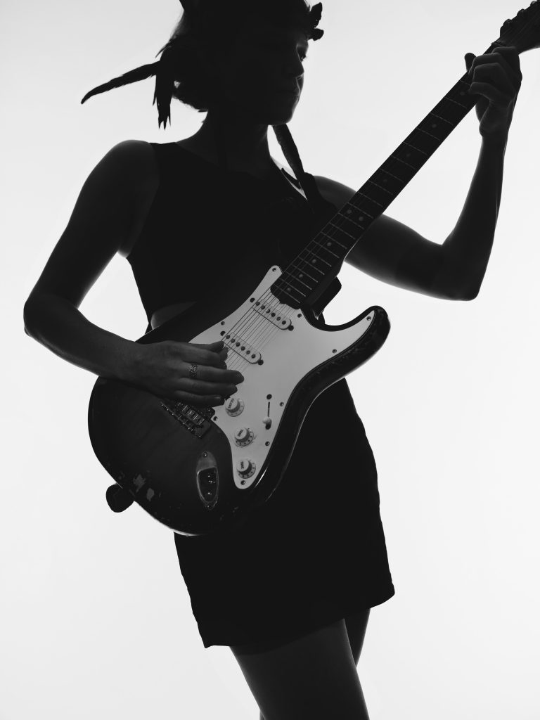 Emanuel Klempa, Fotograf München. Eine schwarz-weiße Musikerfoto-Silhouette einer Person, die eine E-Gitarre spielt und ein ärmelloses Kleid trägt, vor einem einfachen hellen Hintergrund.