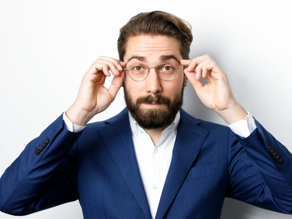 Emanuel Klempa, Fotograf München. Ein Mann mit Bart und Brille, der einen blauen Anzug und ein weißes Hemd trägt, steht vor einem weißen Hintergrund, rückt seine Brille zurecht und blickt im Stil eines professionellen Businessporträts in die Kamera.