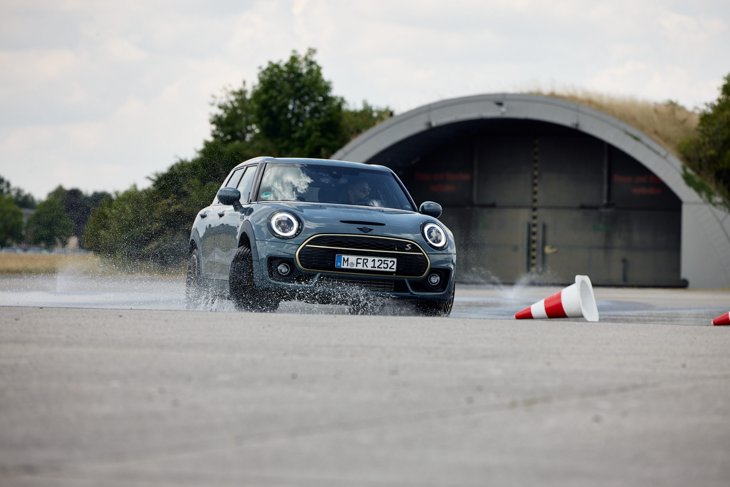 Emanuel Klempa, Fotograf München. Ein dunkler Mini Cooper schlittert bei einem Mini Driving Event in Augsburg über den nassen Asphalt und spritzt an einem umgestoßenen Verkehrsleitkegel vorbei. Im Hintergrund erhebt sich ein großer runder Hangar inmitten üppigen Grüns.