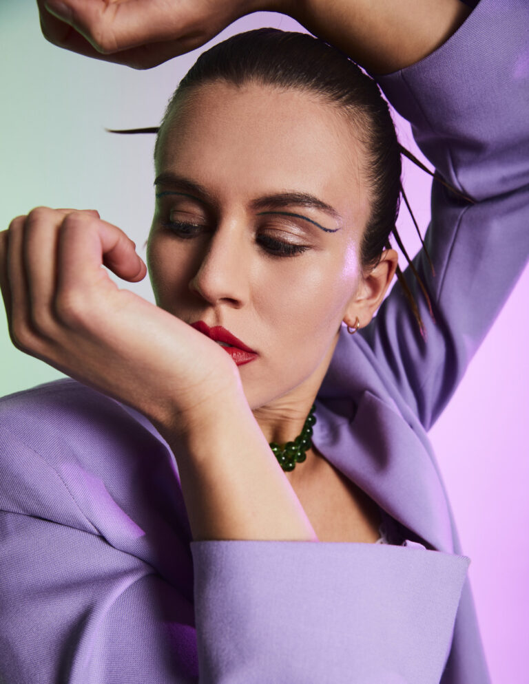 Emanuel Klempa, Fotograf München. Eine Frau in einem hellvioletten Blazer nimmt eine dynamische Pose für ein Mode-Editorial ein, die Arme angehoben und angewinkelt, um ihr Gesicht teilweise zu verdecken. Sie trägt ein auffälliges Make-up, geflügelten Eyeliner, roten Lippenstift und eine grüne Perlenkette vor einem rosa und grünen Hintergrund.