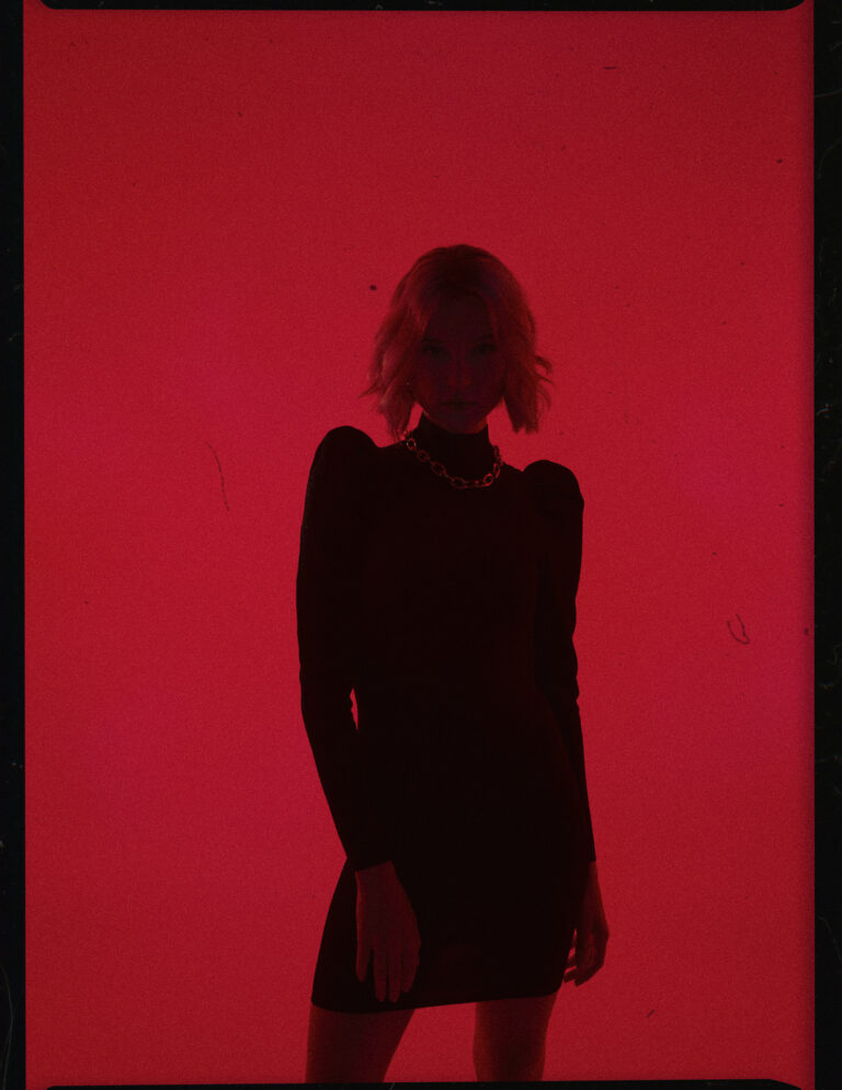 Emanuel Klempa, Fotograf München. Eine Person in einem schwarzen Kleid und mit kurzen Haaren steht vor einem leuchtend roten Hintergrund, wodurch eine auffällige Silhouette entsteht, die an ein High-Fashion Editorial erinnert. Die dramatische Beleuchtung wirft die Figur meist in den Schatten.