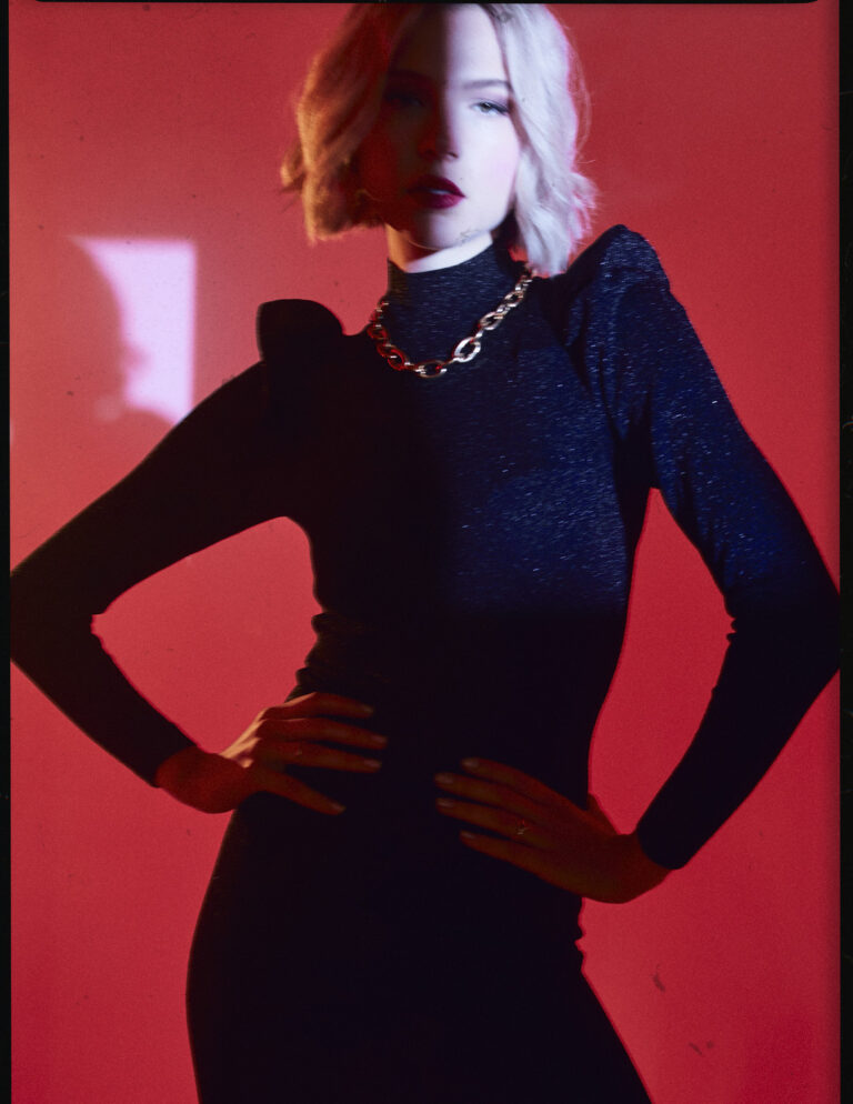 Emanuel Klempa, Fotograf München. Eine Frau mit kurzen blonden Haaren in einem taillierten schwarzen Kleid und einer Kettenhalskette posiert selbstbewusst mit den Händen in den Hüften vor einem leuchtend roten Hintergrund und fängt damit die Essenz eines auffälligen Mode-Editorials ein.