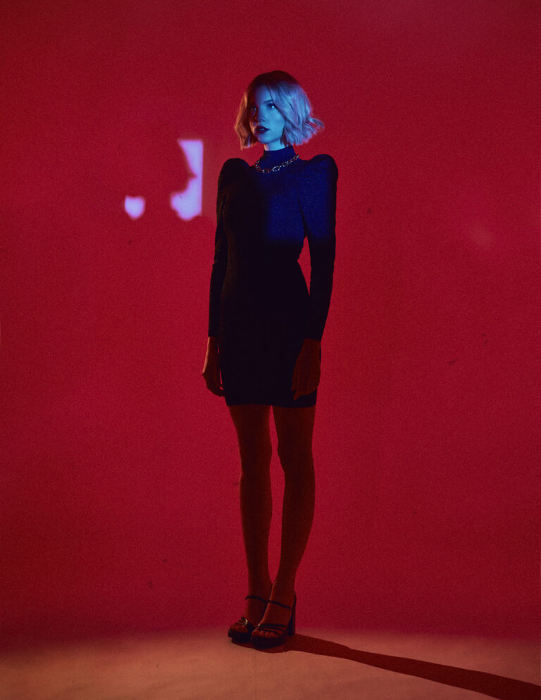 Emanuel Klempa, Fotograf München. Eine Frau in einem schwarzen Kleid und hochhackigen Schuhen steht vor einem leuchtend roten Hintergrund, der mit blauem Licht auf Gesicht und Oberkörper dramatisch beleuchtet wird. Mit kurzen, gewellten blonden Haaren posiert sie für ein auffälliges Fashion Editorial, die Arme entspannt an den Seiten.