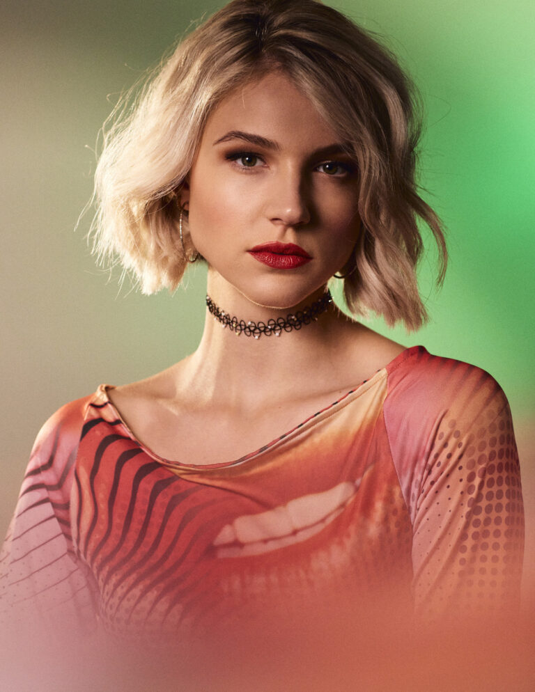 Emanuel Klempa, Fotograf München. Eine Frau mit kurzen blonden Haaren und rotem Lippenstift trägt ein Halsband und ein buntes Hemd mit abstrakten Mustern vor einem unscharfen grünen und beigen Hintergrund und verströmt Fashion Editorial Vibes.