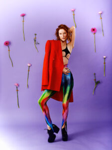 Emanuel Klempa, Fotograf München. In diesem auffälligen Mode-Editorial posiert eine Frau vor einem lila Hintergrund, der mit vertikalen rosa Blumen verziert ist. Sie trägt einen schwarzen BH, regenbogenfarbene Leggings, schwarze Plateauschuhe und drapiert einen roten Mantel über eine Schulter.