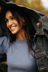 Emanuel Klempa, Fotograf München. Eine lächelnde Frau hält sich eine Jeansjacke über den Kopf, während sie für einen lässigen Modeltest im Freien steht und einen hellblauen Pullover trägt. Der sanft verschwommene Hintergrund zeigt einen Hauch von Herbstfarben und macht diese Aufnahme perfekt für ein Gosee-Portfolio.
