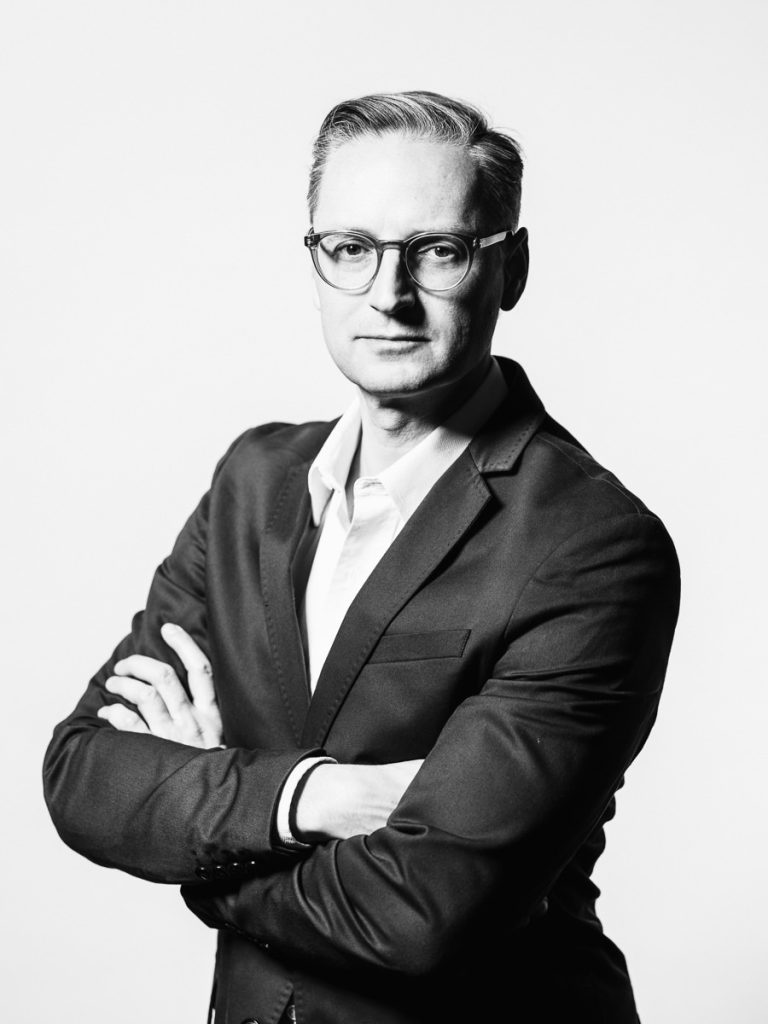 Emanuel Klempa, Fotograf München. Schwarz-Weiß-Foto eines Mannes im Anzug, mit Brille und kurzen Haaren, der mit verschränkten Armen in die Kamera schaut, vor einem schlichten Hintergrund. Ideal für alle, die sich von einem Businessfotografen München inspirieren lassen wollen.