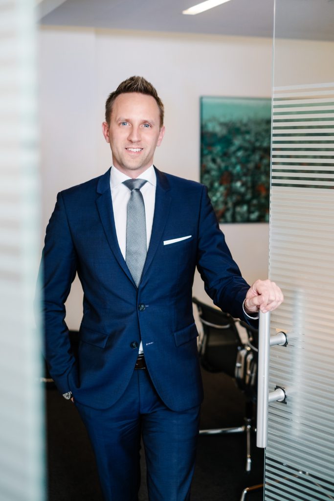 Emanuel Klempa, Fotograf München. Ein Mann in blauem Anzug und grauer Krawatte steht lächelnd am Eingang eines modernen Büros und hält eine Glastür offen. Aufgenommen von Businessfotograf München sind im Hintergrund ein Konferenztisch und Stühle zu sehen.