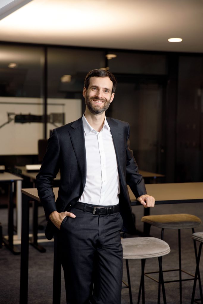 Emanuel Klempa, Fotograf München. Ein Mann in einem dunklen Anzug und weißem Hemd steht lächelnd in einem modernen Büroraum und posiert für Geschäftsporträts, während er sich mit einer Hand auf einen hohen Tisch stützt und die andere in die Tasche steckt. Im Hintergrund sind hohe Stühle und Glaswände zu sehen.